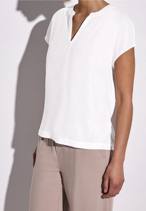 Personne portant un chemisier blanc à manches courtes avec une encolure en V et un pantalon beige, debout devant un fond clair uni.