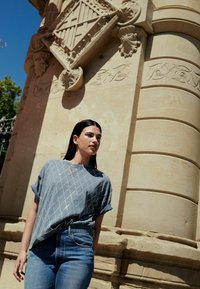 Světle modrý, oversized top s krátkým rukávem, zdobený diamantovým vzorem a s ohrnutými manžetami, spárovaný s tmavě modrými jeansy s vysokým pasem na pozadí z kamene.