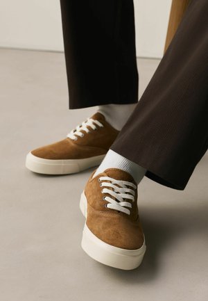 Braune Wildleder-Sneaker mit weißen Schnürsenkeln und dicken weißen Sohlen, getragen mit schwarzen Hosen und weißen Socken auf einem hellen Boden.