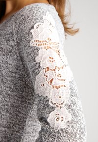 Grå stickad tröja med vit blommig spetsdetalj på vänster ärm; mjuk textur och avslappnad passform; rund halsringning.
