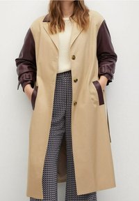 Cappotto beige con maniche in pelle marrone, bottoni dorati e tasche laterali. Indossato sopra un maglione color crema e pantaloni a fantasia.