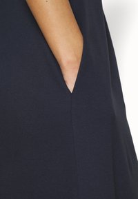 Main d'une personne dans la poche latérale d'un vêtement bleu marine foncé, montrant un gros plan du bras et de la texture du tissu.