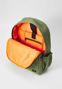 Zaino in tela verde con un design curvo; presenta una fodera interna arancione, una tasca frontale con zip e un pannello superiore imbottito.