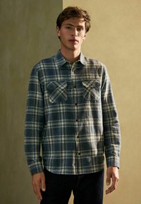Camisa de franela en patrón a cuadros azul y gris, con dos bolsillos en el pecho, diseño de botones y mangas largas con puños abotonados.