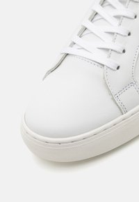 Weißer Leder-Sneaker mit glattem Obermaterial, flachen weißen Schnürsenkeln und einer strukturierten Gummisohle mit erhöhtem Rand. Schlichtes Design mit subtilen Nähten.