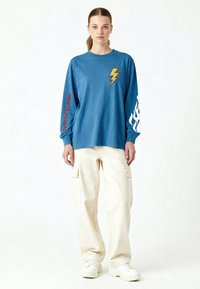 Femme debout portant une chemise bleu à manches longues avec un motif d'éclair et de flammes, un pantalon cargo crème et des baskets blanches à plateforme.