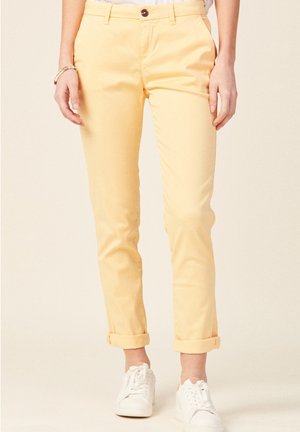 Chinos en coton jaunes avec des revers, dotés d'une fermeture à bouton à l'avant, de poches latérales et d'une texture lisse. Associés à des baskets blanches.
