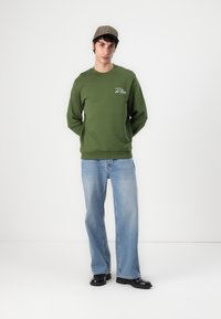 Grüne Sweatshirt mit Logo, lockere Passform, runder Halsausschnitt. Kombiniert mit hellblauen geraden Jeans und schwarzen Schuhen. Eine karierte Mütze rundet den Look ab.