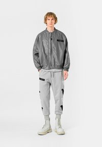 Cazadora bomber de imitación cuero gris con cremallera, combinada con pantalones de chándal gris claro con acentos negros y bordados florales; llevada con botas blancas.