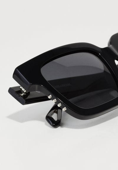 Occhiali da sole neri con montatura quadrata, finitura opaca, lenti scure e dettagli argentati sui lati. Include un design pieghevole unico.