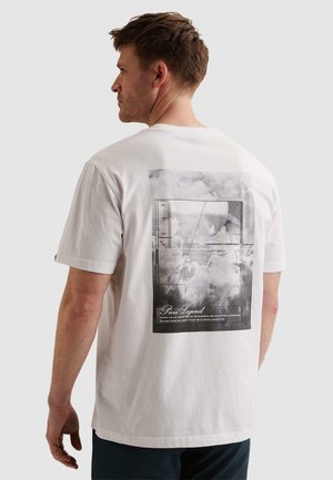 Muž s krátkymi hnedými vlasmi, otočený vľavo, má na sebe biele tričko s čierno-bielou grafikou oblakov a textom na chrbte.