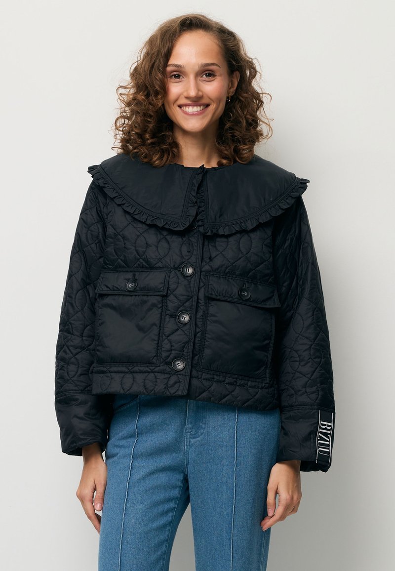 Glimlachende vrouw met krullend haar, gekleed in een zwarte doorgestikte jas met een grote kraag en een blauwe spijkerbroek, staand tegen een effen lichte achtergrond.