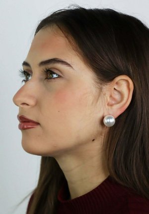 Femme aux cheveux longs et bruns, portant de grandes boucles d'oreilles rondes en perles et un haut rouge foncé, regardant vers la gauche sur un fond uni.
