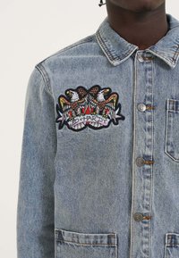 Vaaleansininen farkkutakki klassisella kauluksella, metallinapeilla ja brodeeratulla graafikuvalla, jossa on kotkia ja teksti "ED HARDY."