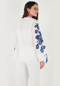 Blusa bianca con ricami floreali blu sulle maniche, caratterizzata da una vestibilità ampia, scollatura rotonda e dettagli con pon pon ai polsini.