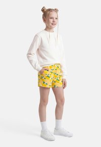 Fille aux cheveux blonds coiffés en deux chignons tressés, portant un sweat-shirt blanc, un short imprimé de fruits jaunes, des chaussettes blanches et des baskets blanches, debout.