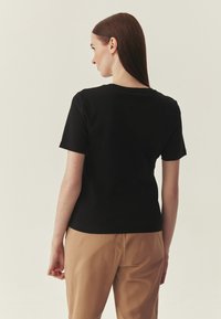 T-shirt noir à manches courtes en tissu de coton lisse, avec un col rond et une coupe ajustée, porté avec un pantalon beige.