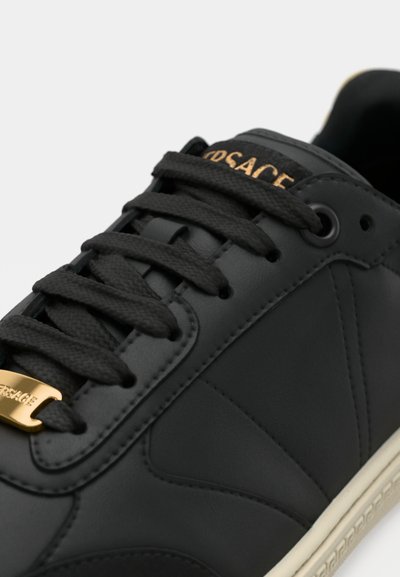 Czarny skórzany sneaker z fakturowaną powierzchnią, ozdobiony złotym logo, okrągłymi sznurowadłami oraz kremową gumową podeszwą. Gładkie wykończenie i minimalistyczny design.