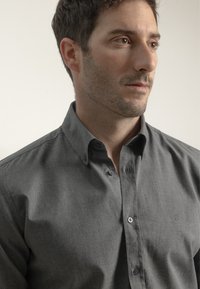 Camisa de botones en tejido gris oscuro. Presenta un cuello clásico, botones en el frente y una textura sutil. Diseño simple con acentos mínimos.