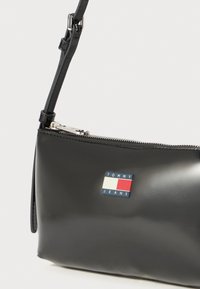 Black glossy handbag with a zip closure, adjustable strap, and a rectangular logo patch in red, white, and blue on the front.