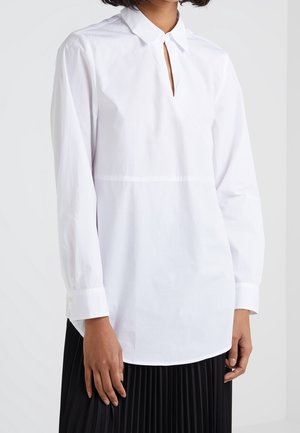 Femme portant un chemisier blanc à manches longues avec une petite fente à l'encolure devant et une jupe plissée noire.