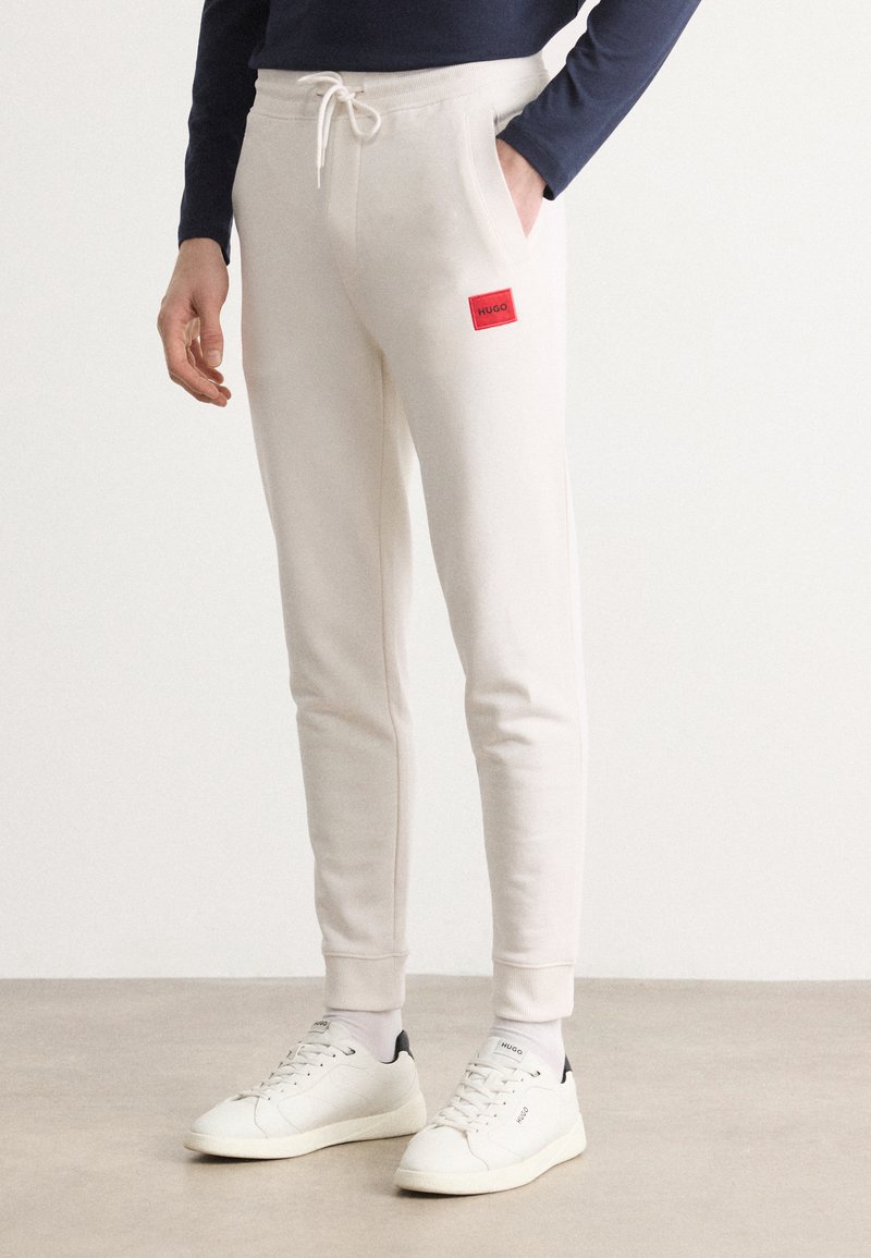 Pantalones deportivos blancos con un diseño ajustado, cinturilla elástica y cordón. Presenta un parche de logo rectangular rojo en la cadera izquierda.