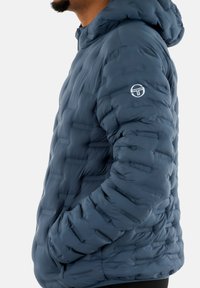 Giubbotto imbottito di colore blu scuro, con texture trapuntata, cappuccio e tasche laterali. Fodera con patch logo sulla manica sinistra. Tessuto liscio, design leggero.