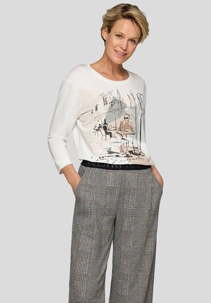 Lächelnde Frau mit kurzen blonden Haaren, die ein weißes Langarmshirt mit Café-Illustration und karierten, weit geschnittenen Hosen trägt.