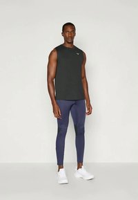 Fekete ujj nélküli sportpólót, sötétkék, testhez simuló leggingset fekete foltokkal és fehér futócipőt viselő férfi, sima háttér előtt áll.