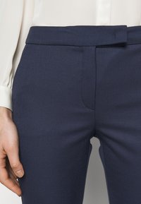 Granatowe, dopasowane spodnie uszyte z teksturowanej tkaniny, z gładkim przodem i gładkim paskiem w talii. Posiadają boczne kieszenie i wąski krój.