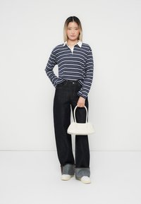 Marineblå og hvite stripete langermet polo skjorte, mørke vide jeans, hvit handveske og sneakers. Modellen står mot en ensfarget bakgrunn.
