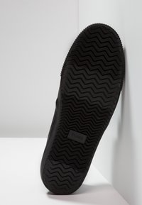 Musta kumipohja, jossa on sahalaitakuvio, varustettuna suorakaiteen muotoisella logotarralla, jossa on kohokuvioituna "TOMS". Pinta on teksturoitu ja sileä.