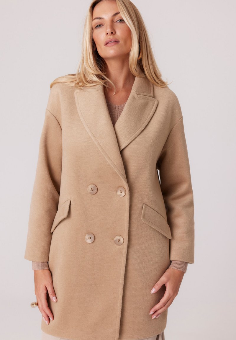 Beżowy płaszcz dwurzędowy z kieszeniami z przodu, klapami i subtelną fakturą. Posiada duże guziki i akcenty na mankietach w jaśniejszym odcieniu.