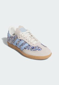 Hellgrauer Sneaker mit blauem Blumenmuster, aus Wildleder, drei blauen Streifen und einer Sohle aus Gummi. Zackenförmiges Zungen-Design.
