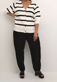 Valko-mustaraidallinen neuletakki, jossa V-kaula-aukko ja napitettu kiinnitys, yhdistetty mustiin leveälahkeisiin housuihin ja mustiin avonaisiin korkokenkiin.