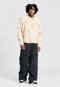 Pullover beige morbido con colletto e vestibilità comoda, abbinato a pantaloni cargo neri con molteplici tasche e taglio ampio.