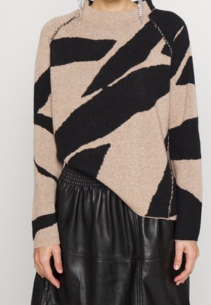 Pull beige et noir à motif abstrait associé à une jupe en cuir noire froncée, porté par une personne avec des boucles d’oreilles pendantes.