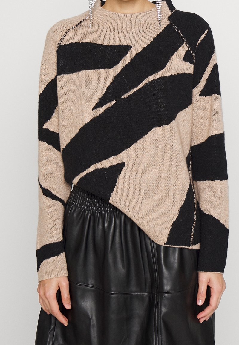 Pull beige et noir à motif abstrait associé à une jupe en cuir noire froncée, porté par une personne avec des boucles d’oreilles pendantes.