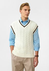 Witte gebreide vest met kabelpatroon, voorzien van een donkerbruine V-halsbies. Draagt over een lichtblauw gestreept shirt en beige broeken.