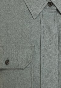 Camicia grigia con bottoni, realizzata in tessuto testurizzato, colletto a punta e taschino sul petto. Presenta bottoni scuri e dettagli di cucitura in vista.