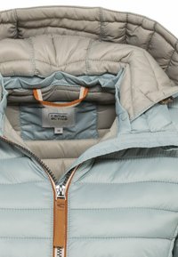 Chaqueta acolchada de color azul claro con capucha de tipo acolchado en beige, tirador de cremallera marrón y etiqueta de la marca Camel Active visible en el interior del cuello.