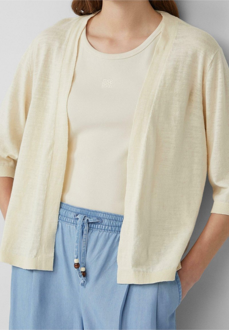Femme portant un cardigan crème à manches courtes sur un top crème et un pantalon bleu clair à cordon avec des perles sur le cordon.