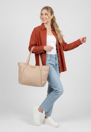 Jauna moteris su ilgais šviesiais plaukais dėvi rūdžių spalvos marškinius, baltą palaidinę, šviesius džinsus, baltus sportinius batelius ir ant peties neša didelę smėlio spalvos rankinę.