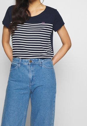 Femme portant un t-shirt rayé noir et blanc à manches courtes avec petit détail cœur rose et un jean bleu taille haute, les mains dans les poches.
