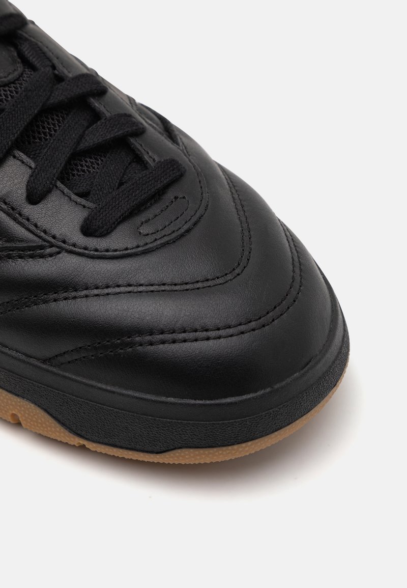 Zapatilla de deporte negra de cuero con textura suave, punta redonda, detalles perforados, cordones negros y suela de goma color caramelo.