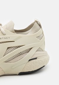 Närbild av en beige sportskor med meshtyg, krämfärgade strukturoverlägg, vadderad sula och svart greppdetalj på yttersulan.