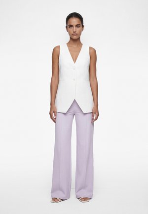 Frau trägt eine ärmellose weiße Weste mit Knöpfen und weit geschnittene hellviolette Hosen, steht barfuß vor einem weißen Hintergrund.
