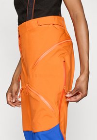 Pantaloni outdoor arancioni con multiple tasche con cerniera angolare, vita alta e una sezione inferiore blu a contrasto con dettagli testurizzati.