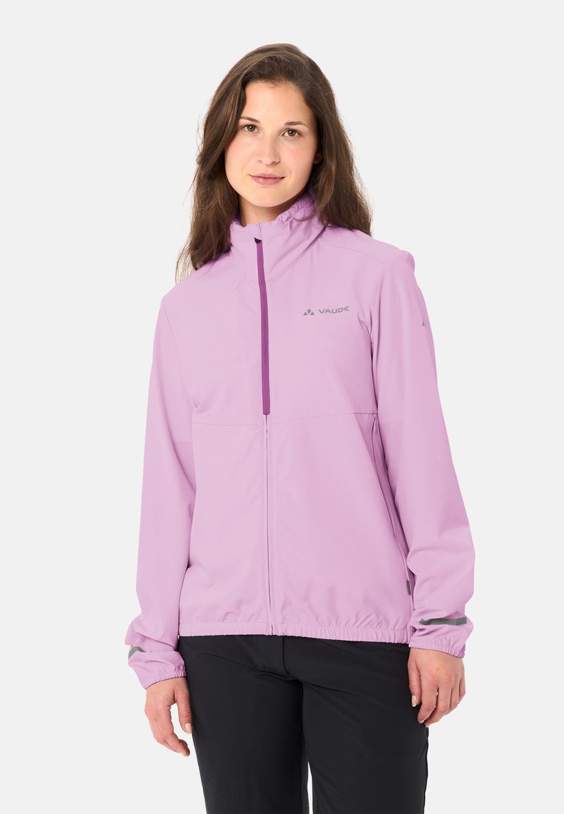 Vrouw die een lichtpaarse Vaude-jas met ritssluiting en een zwarte broek draagt, staand tegen een effen lichte achtergrond.