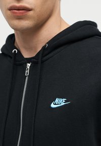 Νεαρό άτομο που φορά μαύρο φούτερ Nike με φερμουάρ και ανοιχτό μπλε λογότυπο στο στήθος, ορατό από τον λαιμό μέχρι το πάνω μέρος του κορμού.